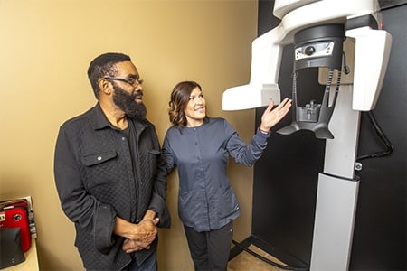 Dental Assistant showing a patient the 3D Cone Beam CT scanner used for planning precision dental implant placement.